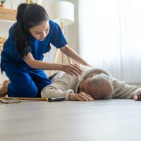 Caregiver helping senior male from falling on the ground at home. Elderly older patient having an accident after doing physical therapy then rescued by attractive therapist nurse in living room.