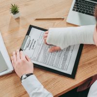 cropped view of worker holding broken arm compensation claim and sitting at table in office.