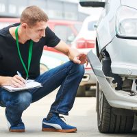 Insurance agent recording damage after car crash during inspecting damaged automobile on claim form