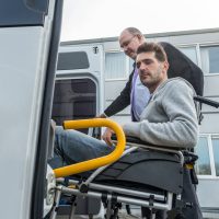 A man in a wheelchair is being assisted into a specialized transport van by an attendant or lawyer outside a modern building.