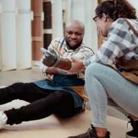 Multiracial foreman injured at leg from accident crying with pain in warehouse. Female latin engineer help colleague who falling down get physical injury in workplace. Diverse people in workplace.