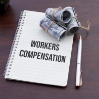A notebook labeled "Workers Compensation" accompanied by cash, symbolizing financial planning and legal considerations, rests on a rustic wooden table.