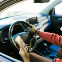 A person in a car holds a smartphone showing text messages, with one hand on the steering wheel, suggesting distracted driving. Bright daylight.