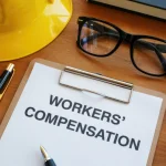 workers-compensation-claim-paperwork-and-protective-hard-hat-resting-on-a-wooden-desk-alongside-reading-glasses-and-writing-instruments